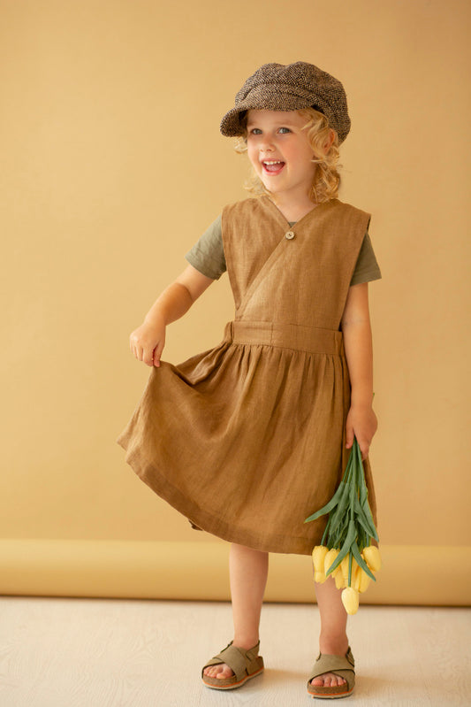 A young child wearing a brown linen-look sleeveless dress over a muted green short-sleeve shirt, a brown textured flat cap, and green sandals, holding a bunch of yellow tulips against a warm beige studio backdrop.