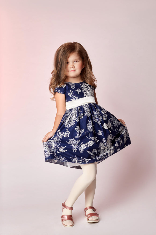 A young girl with wavy light brown hair stands in a studio wearing a navy blue dress with a white toile-style print, a white waistband, white tights, and metallic rose gold sandals against a soft pink background.