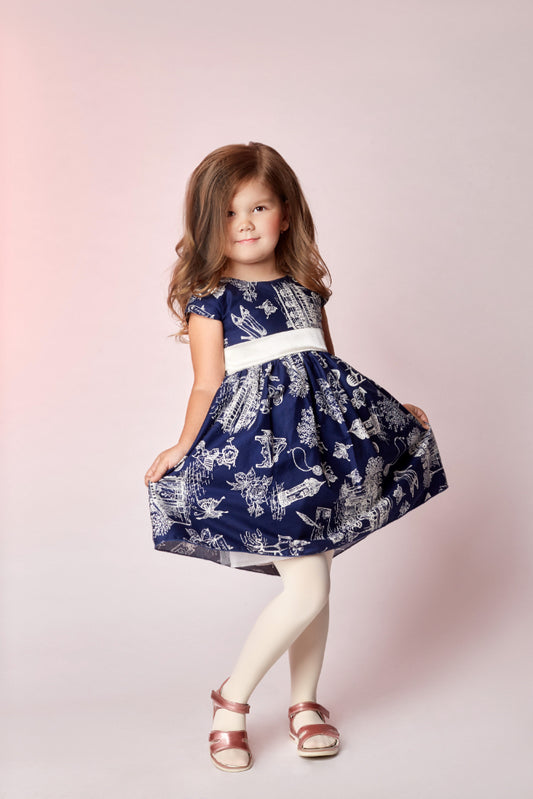 A young girl with wavy light brown hair stands in a studio wearing a navy blue dress with a white toile-style print, a white waistband, white tights, and metallic rose gold sandals against a soft pink background.