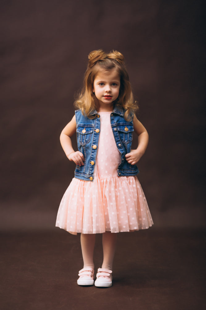 A young girl stands against a dark brown studio backdrop wearing a light pink dress with a white polka-dot tulle skirt, a blue denim vest, and white shoes.